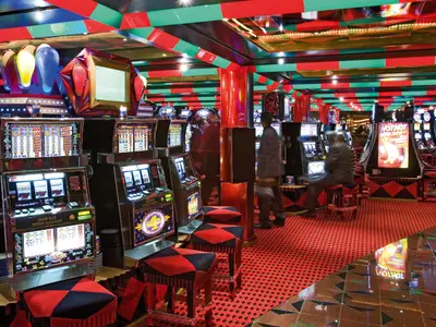 Spielautomaten und rote Sitzhocker im Casino Giano an Bord der Costa Serena mit Gästen beim Spielen.