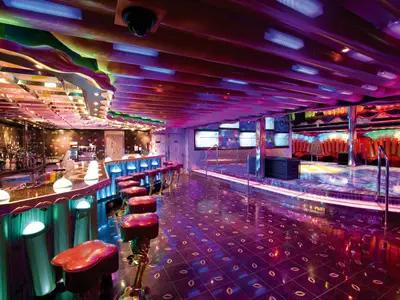 Die farbenfroh beleuchtete Diskothek an Bord der Costa Serena zeigt eine Bar mit roten Barhockern, eine Tanzfläche und eine Lichtinstallation.