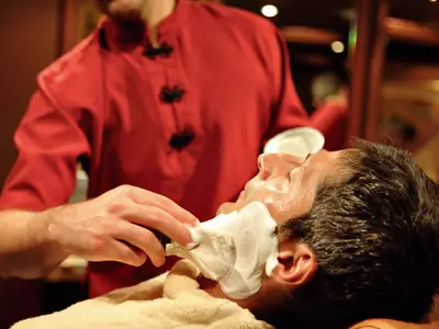 Ein Barbier trägt Rasierschaum auf das Gesicht eines entspannten Mannes im Spa-Bereich an Bord der Costa Serena auf.