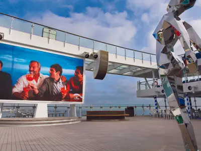 Blick auf das sonnige Außendeck der Costa Smeralda mit einer großen LED-Leinwand und einer glänzenden, futuristischen Metallskulptur.