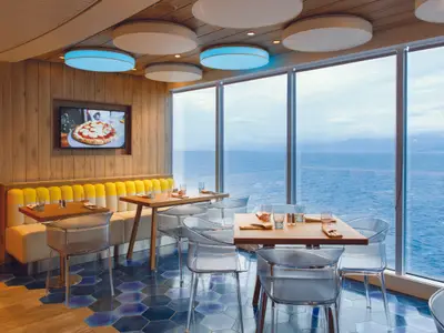 Gedeckte Tische mit transparenten Stühlen im Restaurant Tutti a Tavola an Bord der Costa Smeralda vor großen Fenstern mit Blick aufs Meer.