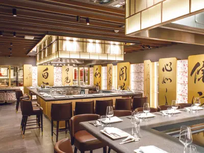 Das Teppanyaki-Restaurant an Bord der Costa Smeralda mit eleganten Barhockern, goldenen Wänden mit Schriftzeichen und gedeckten Tischen.