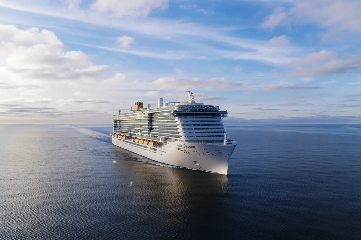 Die Costa Toscana in einer Luftaufnahme, die das große Kreuzfahrtschiff von vorne-schräg zeigt, wie es bei Tageslicht auf dem offenen Meer fährt. Der Himmel ist teilweise bewölkt.
