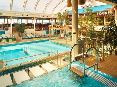 Der lichtdurchflutete Innenpool "La Spiaggia Beach Club" an Bord der Costa Toscana mit Liegestühlen, Pflanzen und einem Whirlpool im Vordergrund.