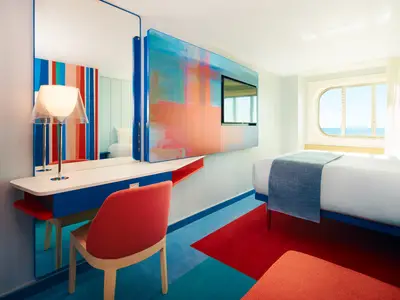 Helle Aussenkabine an Bord der Costa Toscana mit modernem, farbenfrohem Design. Ein Bett, Schreibtisch, Stuhl und ovales Fenster mit Meerblick sind zu sehen. Blau, Rot und Türkis dominieren die Einrichtung.