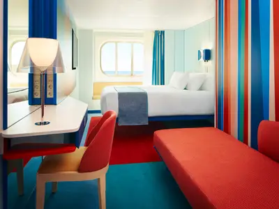 Geräumige Aussenkabine an Bord der Costa Toscana mit farbenfrohem Design, Schreibtisch, Bett und einem runden Fenster mit Meerblick. Roter Teppich und Sofa.