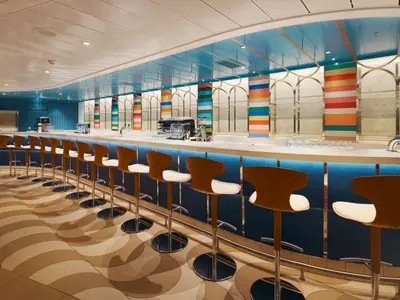 Eine lange, moderne Bar an Bord der Costa Toscana mit braun-weißen Barhockern. Die Rückwand ist mit vertikalen Streifen in verschiedenen Farben und goldenen, geschwungenen Mustern verziert. Die Bar ist hell beleuchtet, die Decke ist hellblau.