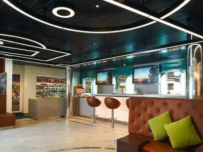 Ein Blick in die moderne Bar an Bord der Costa Toscana mit braunen Ledersofas, grünen Kissen, Barhockern und einer gut bestückten Theke. Überall strahlen weiße Neonlichter, die eine lebendige Atmosphäre schaffen.