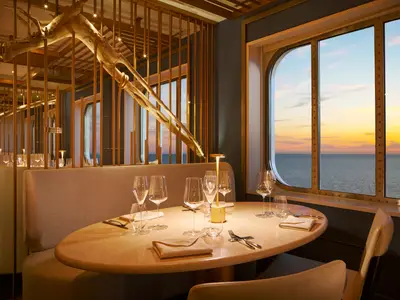 Ein gedeckter runder Tisch im Archipelago Restaurant an Bord der Costa Toscana mit Blick durch das Fenster auf den Sonnenuntergang über dem Meer.