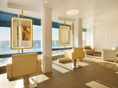 Hell erleuchteter Friseursalon an Bord der Costa Toscana mit beigen Sesseln, großen beleuchteten Spiegeln und Blick auf das weite Meer.