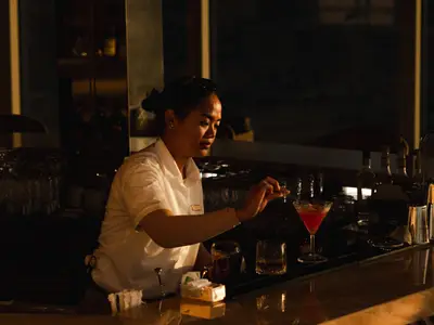 Barkeeperin in weißer Bluse richtet an der stimmungsvoll beleuchteten Bar der Explora I einen roten Cocktail an.
