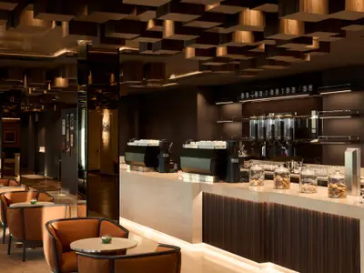 Moderne Kaffeebar an Bord der Explora I mit Kaffeemaschinen, Gebäckgläsern, Holzdecke und stilvollen braunen Sesseln.