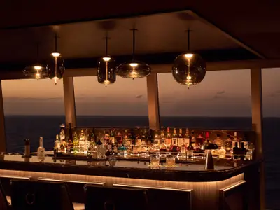 Eine beleuchtete Bar an Bord der Explora II mit Whiskyflaschen, Gläsern und Meerblick bei Sonnenuntergang.