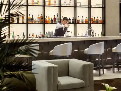 Ein Barkeeper bereitet an einer eleganten Bar mit beleuchteten Regalen und gemütlichen Sesseln an Bord der Explora II einen Cocktail zu.