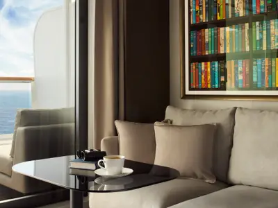 Blick in eine luxuriöse Suite an Bord der Explora II mit Sofa, Kaffeetasse, Buchkunst und Balkon zum Meer.