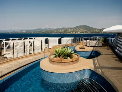 Poolbereich an Bord der Explora III mit blauen Fliesen, Pflanzeninsel, Cabanas und Meerblick auf eine Küstenlandschaft.