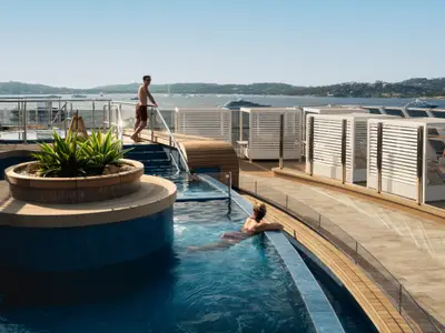 Frau entspannt im glasklaren Wasser des Helios Pools an Bord der Explora III vor mediterraner Kulisse.
