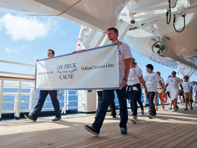 Passagiere und Crew tragen an Bord der Eurodam ein Banner beim 'On Deck for a Cause'-Spendenlauf auf dem Außendeck bei Sonnenschein.