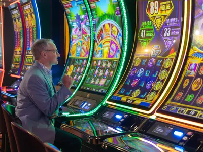 Ein lächelnder Mann mit Brille und Sakko sitzt an Bord der Eurodam vor leuchtenden Spielautomaten im Casino.
