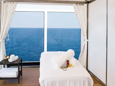 Eine Massageliege mit weißen Handtüchern im Spa-Bereich an Bord der Eurodam, mit Blick durch große Fenster auf das weite, blaue Meer.