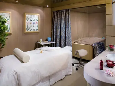 Blick in einen hellen Behandlungsraum im Spa an Bord der Eurodam mit einer Massageliege, einem Therapiebett und Pflegeprodukten.