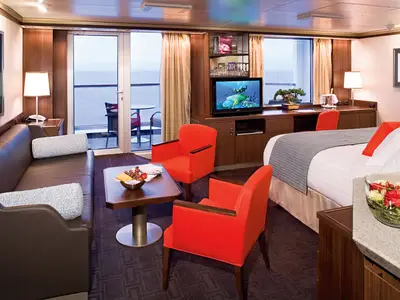 Blick in eine geräumige, barrierefreie Suite an Bord der Eurodam mit zwei roten Sesseln, Sofa, Doppelbett und Balkon.