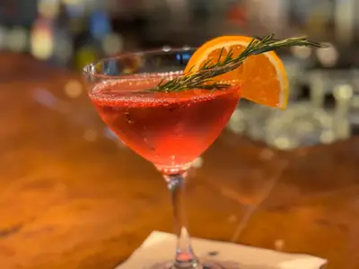 Ein roter Cocktail in einem eleganten Glas, garniert mit einer Orangenscheibe und Rosmarin, serviert an Bord der Koningsdam auf einer Holztheke.