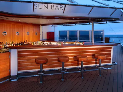 Eine elegante Holzbar mit Barhockern an Bord der Koningsdam bei blauer Stunde mit Blick auf das offene Meer.