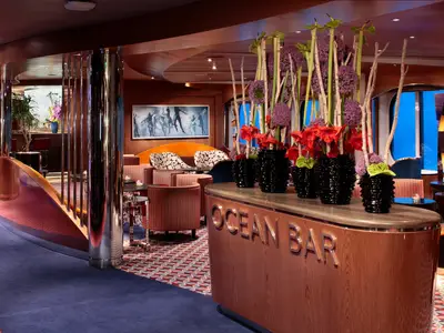 Blick auf die stilvolle Ocean Bar an Bord der Koningsdam mit hölzerner Theke, farbenfrohen Blumengestecken und gemütlichen Sesseln im Hintergrund.
