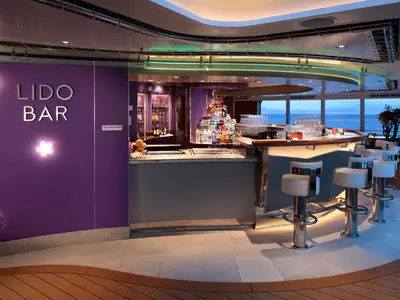 Innenansicht der stilvollen Lido Bar an Bord der Koningsdam mit einer lila Wand, weißen Barhockern und Blick auf das abendliche Meer.