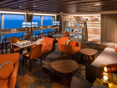 Bibliotheksbereich an Bord der Koningsdam mit gemütlichen Sesseln, Schachspiel und Blick auf das Meer am Abend.