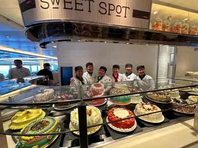 Sechs lächelnde Crew-Mitglieder stehen hinter einer reich bestückten Torten-Vitrine im Buffet-Bereich an Bord der Koningsdam.