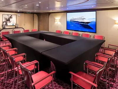 Moderner Konferenzraum Hudson Room an Bord der Koningsdam mit U-förmigem Tisch, roten Stühlen und einem großen Bildschirm.