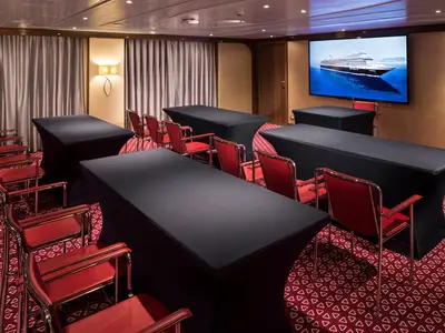 Ein elegant eingerichteter Konferenzraum an Bord der Koningsdam mit schwarzen Tischen und roten Sesseln vor einem Bildschirm.