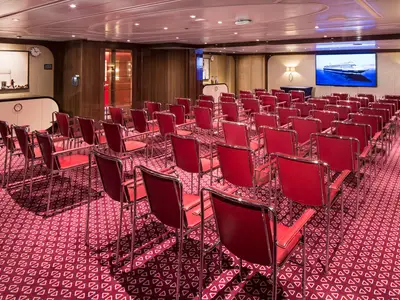 Konferenzraum Hudson Room an Bord der Koningsdam mit Reihen von roten Stühlen vor einem Bildschirm.