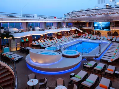 Blick auf den beleuchteten Lido-Pool mit Whirlpools und Liegestühlen an Bord der Koningsdam bei Dämmerung.
