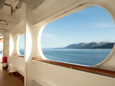 Blick durch ein großes ovales Decksfenster an Bord der Koningsdam auf das ruhige Meer und die Berge Alaskas unter blauem Himmel.