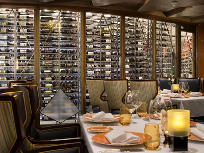 Elegant gedeckte Tische und gestreifte Sessel im Pinnacle Grill Restaurant an Bord der Koningsdam vor einer großen Wand aus Weinregalen.