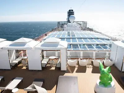 Exklusiver Außenbereich 'The Retreat' an Bord der Koningsdam mit privaten Cabanas, Sesseln, einer grünen Hasen-Skulptur und Blick aufs Meer.