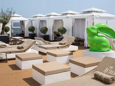 Sonnendeck-Bereich 'The Retreat' an Bord der Koningsdam mit beigen Liegen, weißen Cabanas und einer großen grünen Hasenskulptur.