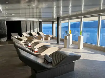 Ruheraum an Bord der Koningsdam mit gemütlichen Wellness-Liegen und großen Panoramafenstern mit Blick auf das Meer und schneebedeckte Berge.