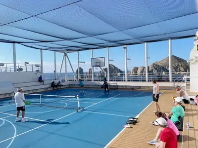 Passagiere spielen Pickleball auf dem Sportdeck an Bord der Koningsdam, während im Hintergrund markante Felsformationen im Meer aufragen.