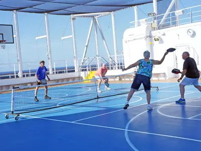 Vier Passagiere spielen aktiv Pickleball auf dem blauen Außensportplatz an Bord der Koningsdam unter blauem Himmel.
