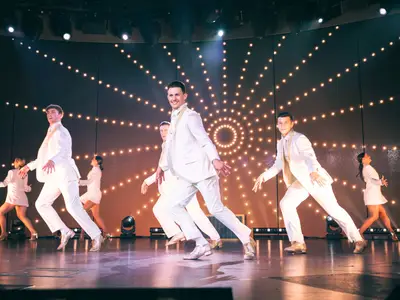 Tänzerinnen und Tänzer in weißen Outfits performen auf der hell erleuchteten Bühne an Bord der Koningsdam.