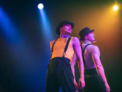 Zwei Tänzer mit Hüten und Hosenträgern bei einer Show auf der Bühne der World Stage an Bord der Koningsdam unter farbigem Scheinwerferlicht.