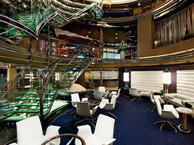 Blick auf das stilvolle, mehrstöckige Atrium an Bord der Nieuw Amsterdam mit einer Treppe aus Glas und gemütlichen Sitzbereichen.