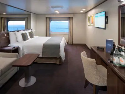 Blick in eine elegante Außenkabine mit Doppelbett, Sitzecke, Schreibtisch und großem Fenster mit Meerblick an Bord der Nieuw Amsterdam.
