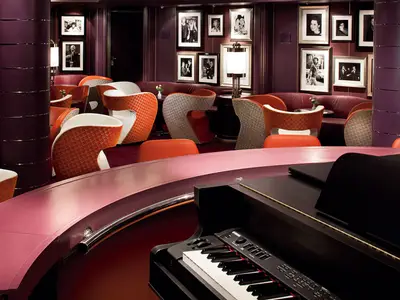 Blick über die Tasten eines Klaviers auf die stilvolle Bar-Lounge mit bunten Sesseln an Bord der Nieuw Amsterdam.