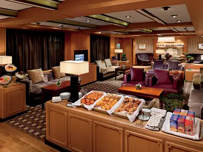 Die Neptune Lounge an Bord der Nieuw Amsterdam mit gemütlichen Sofas, warmem Licht und einer Auswahl an Gebäck auf einem Buffet.