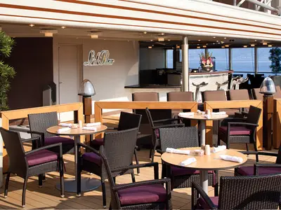 Gedeckte Holztische und dunkle Sessel an Deck der Lido Bar an Bord der Nieuw Amsterdam mit Blick aufs blaue Meer im Hintergrund.